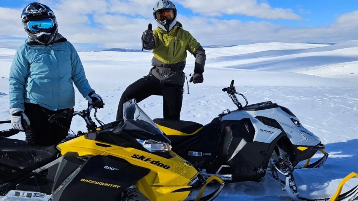Two people pose with snowmobiles on a snowy landscape under a blue sky.