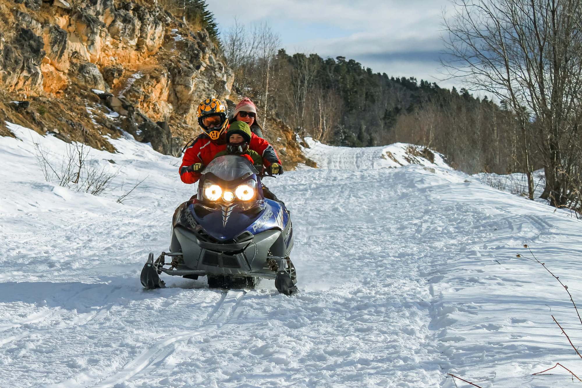 A family rides a snowmobile with the lights on. In the background are rocks, woods and cloudy skies