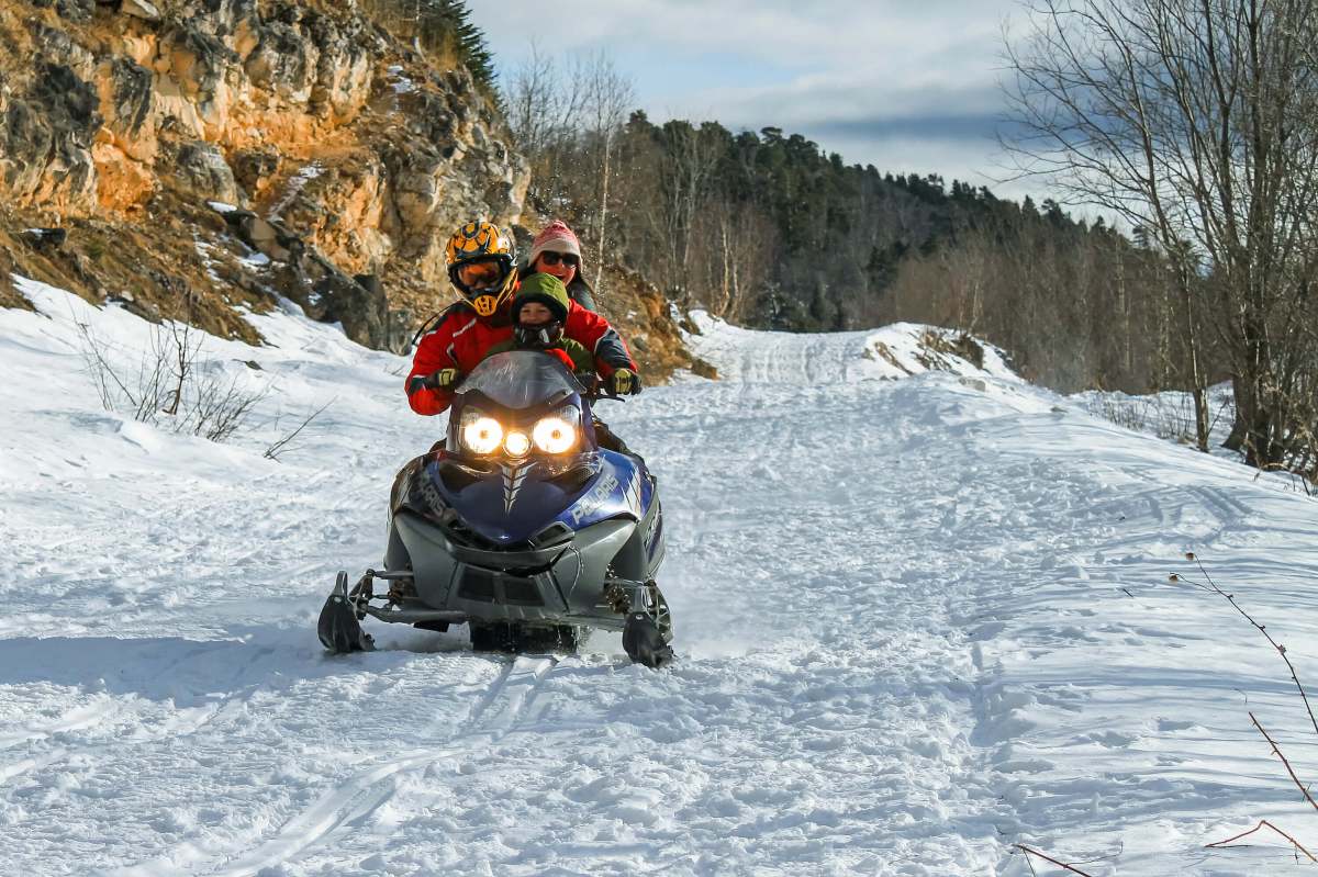 iStock-2161072576 (1) A family rides a snowmobile with the lights on. In the background are rocks, woods and cloudy skies