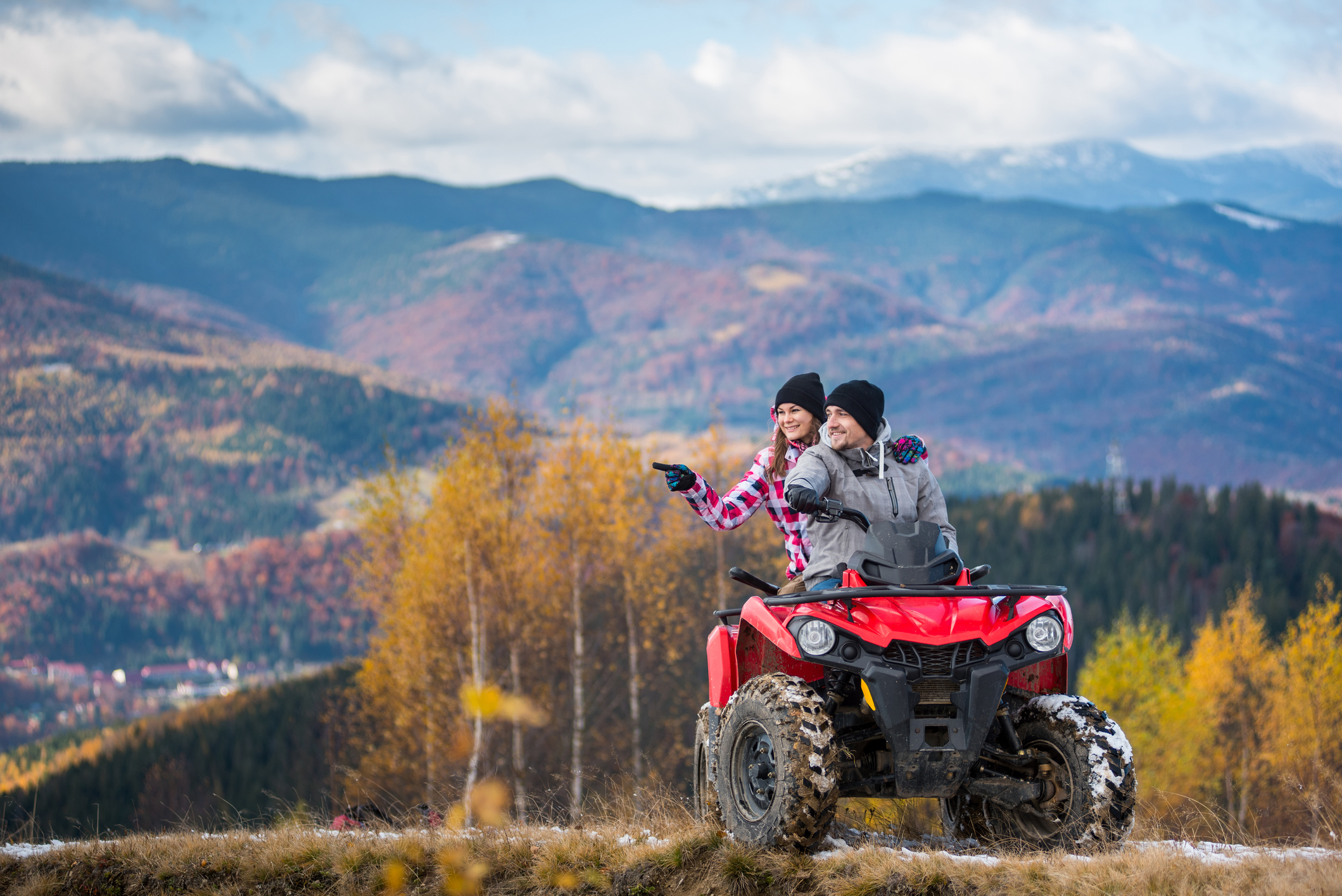 Happy couple on red quad bike riding in the mountains. Young man driving the ATV, girlfriend pointing at something interesting. Beautiful landscape of mountains and forest at autumn day Happy couple on red quad bike riding in the mountains. Young man driving the ATV, girlfriend pointing at something interesting