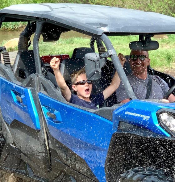 rf Child and adult riding in a blue ATV, splashing through water with arms raised in excitement.