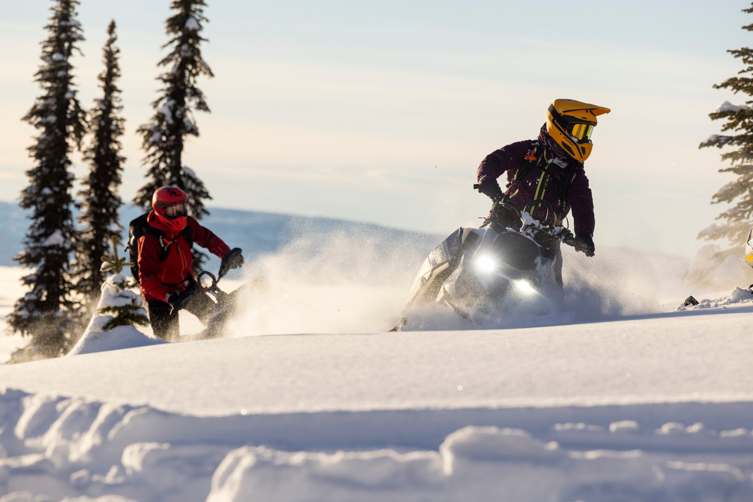 Two people snowmobiling through fresh powder in Utah at sunset, with snow-covered terrain glowing under warm, colorful skies.
