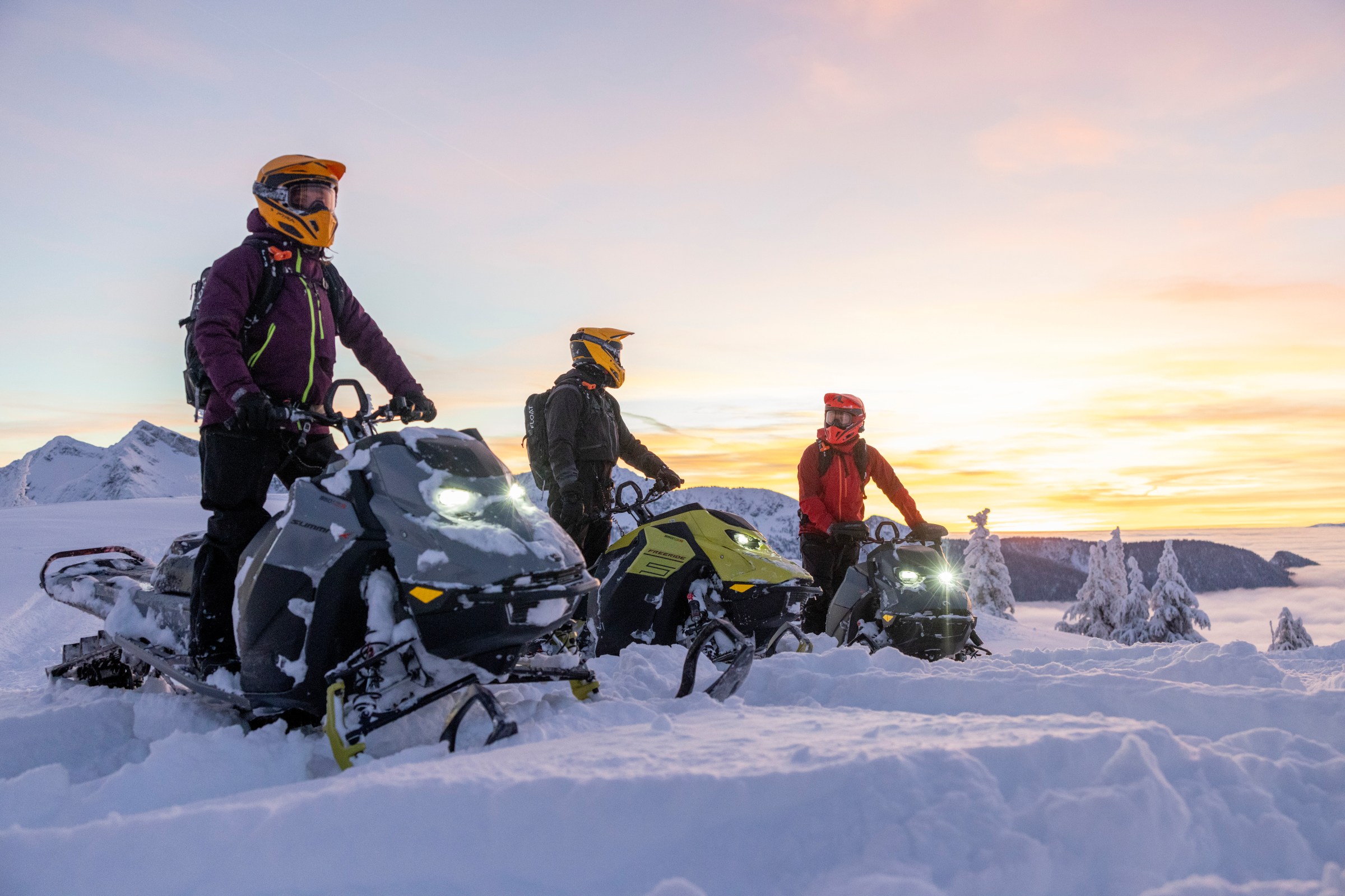 Guided group of three people snowmobiling through fresh snow, following a trail across a snowy landscape with surrounding mountains.