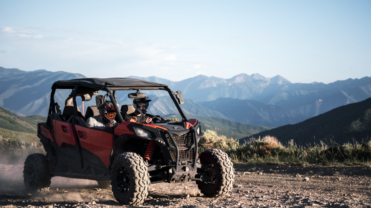 Two people in a red off-road vehicle on a dirt trail with mountains in the background.