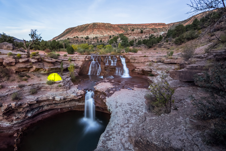 A yellow-green tent glowing from within sits on a rocky cliff in Utah, overlooking cascading waterfalls that flow into a larger waterfall below, surrounded by rugged canyonlands.